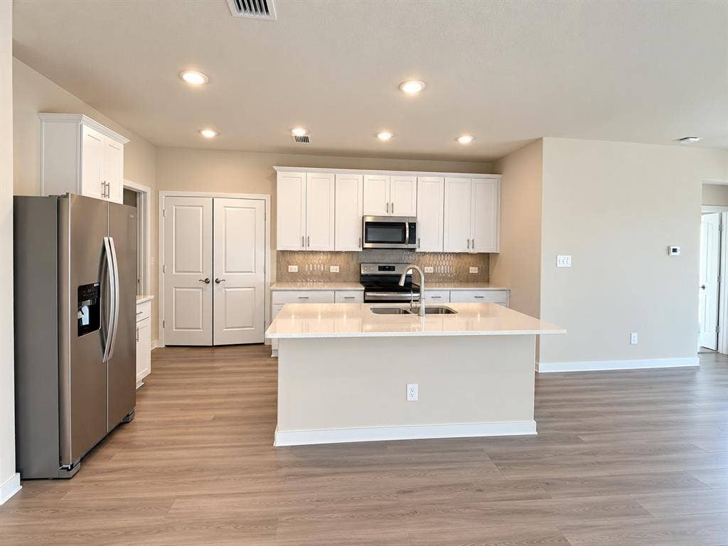 Kitchen featuring white cabinetry, quartz countertops, a central island with a double-basin sink, stainless steel appliances, and wood-finish flooring