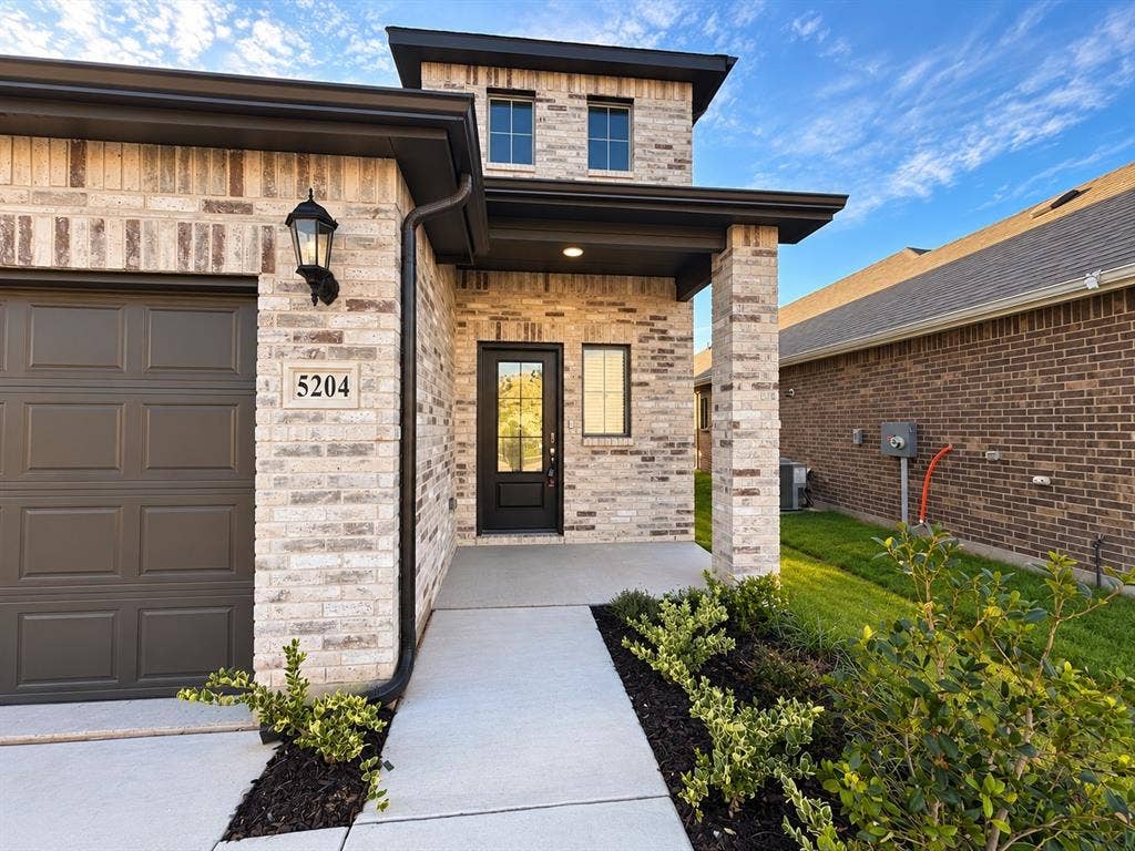 Multi-level exterior featuring light-toned brick masonry, dark metal trim, recessed lighting, and a paved concrete walkway