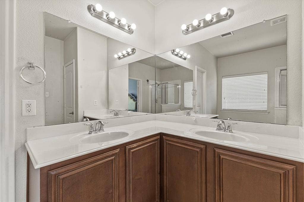 Corner vanity featuring wood-finish cabinetry, dual undermount sinks, solid surface countertops, polished chrome fixtures, and wall-to-wall mirrors with integrated multi-bulb lighting