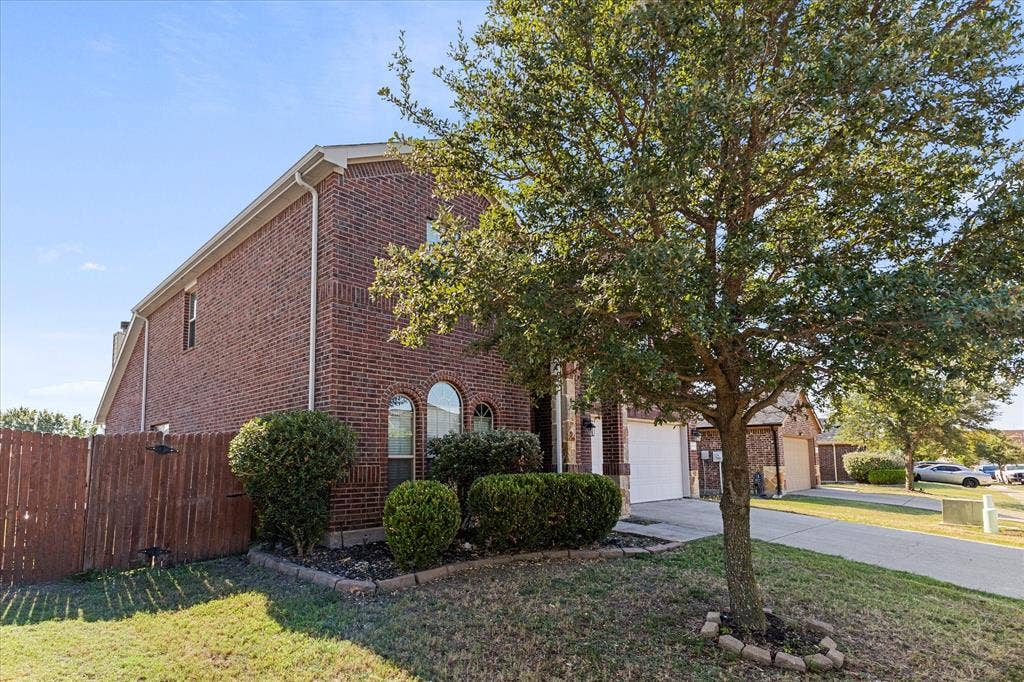Two-story brick exterior featuring arched windows, a front-facing attached garage, and landscaped garden beds with mature trees