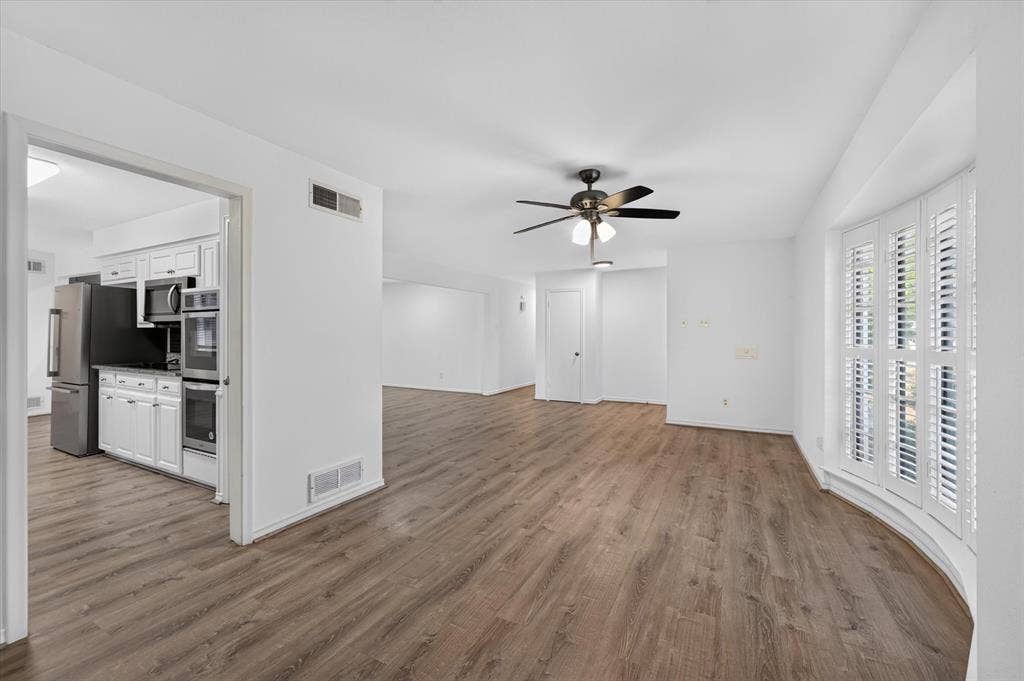 Open-concept interior featuring wood-finish flooring, a bay window with louvered shutters, and a ceiling fan with integrated lighting