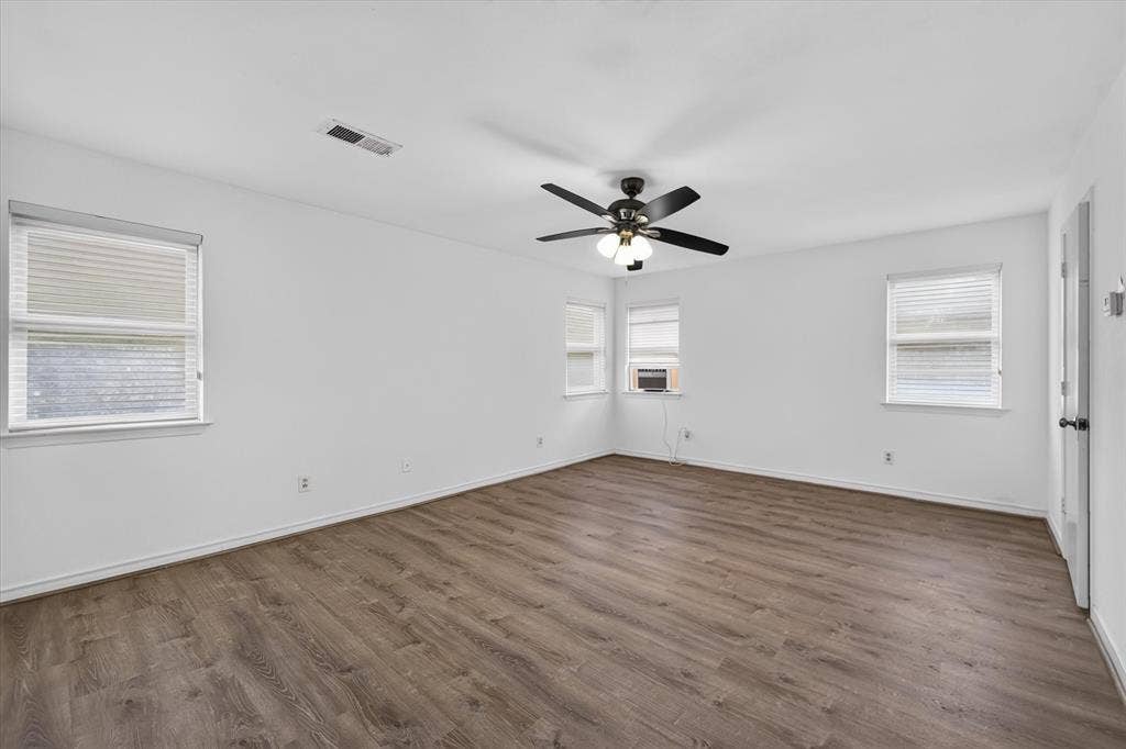 Interior room featuring wood-finish flooring, a neutral color palette, multiple windows with blinds, and a multi-blade ceiling fan with integrated lighting