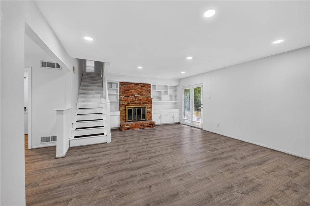 Open living area featuring wood-finish flooring, recessed lighting, red brick fireplace, built-in cabinetry, and double glass doors