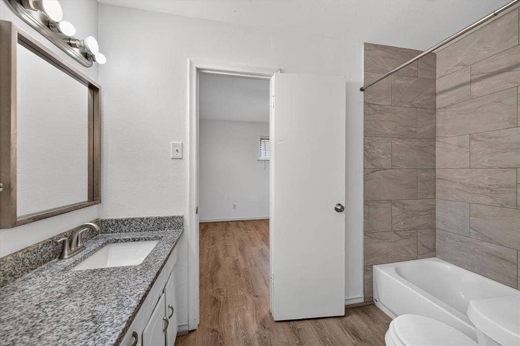 Bathroom featuring a stone-look countertop with an integrated basin, wooden-frame mirror, multi-bulb vanity light, and a shower area with horizontal neutral-toned tile surround