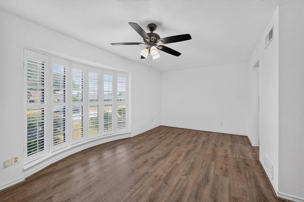 Room featuring wood-finish flooring, a bay window with white plantation shutters, and a central ceiling fan with integrated lighting