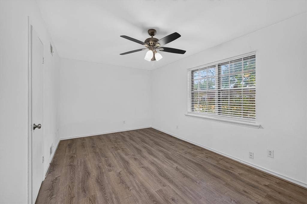Neutral interior featuring wood-finish flooring, a multi-blade ceiling fan with integrated lighting, and a window with horizontal blinds
