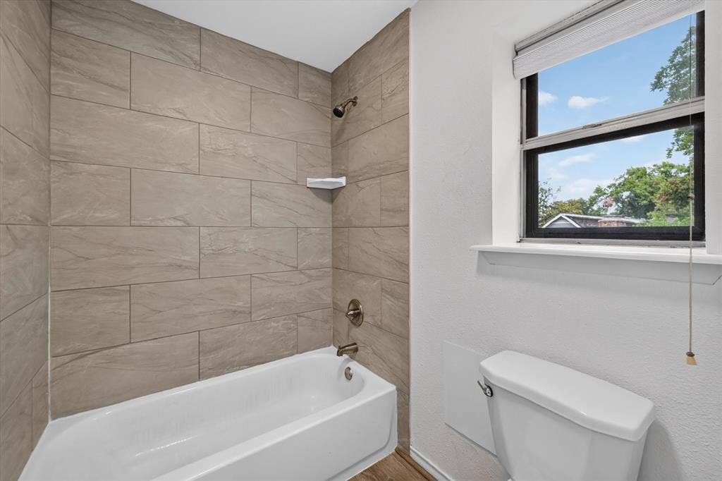 Bathroom featuring a tub-shower combination with floor-to-ceiling large-format tile surround, integrated corner shelving, bronze-finish hardware, and a single-hung window