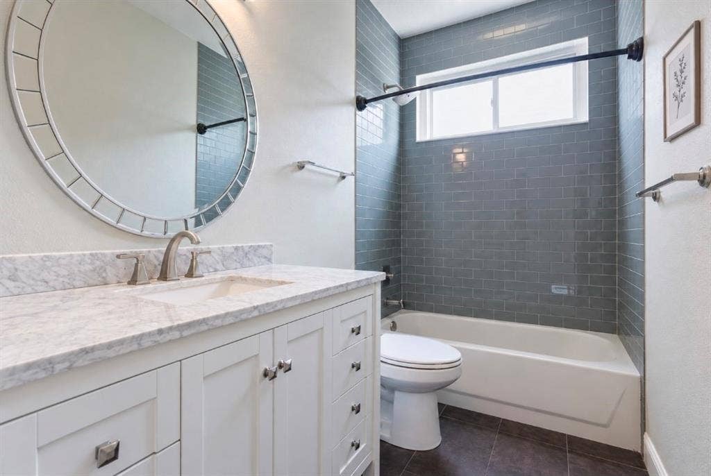 Vanity with stone-finish countertop, undermount sink, and polished nickel faucet, featuring a large circular wall mirror, grey subway tile tub surround, and dark tiled flooring