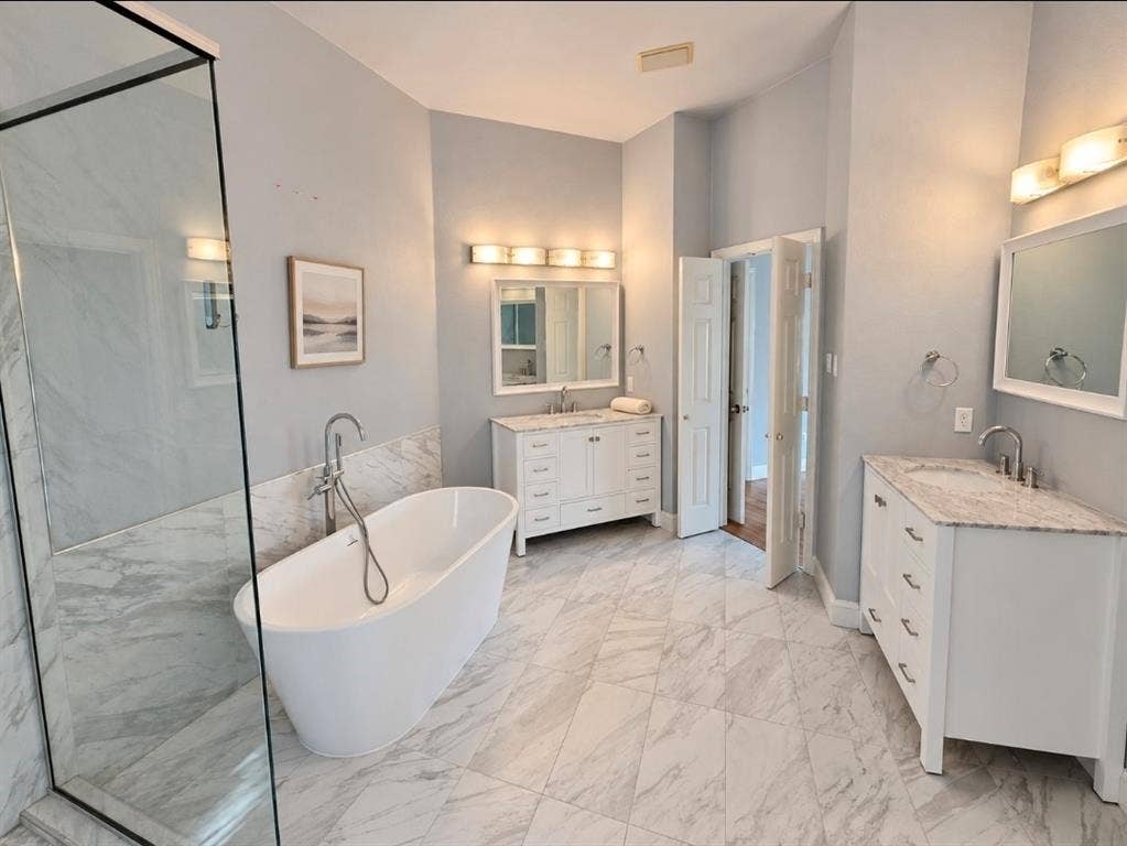 Freestanding soaking tub with chrome floor-mounted faucet, dual white vanities with stone-style countertops, frameless glass-enclosed shower, and large-format tile flooring