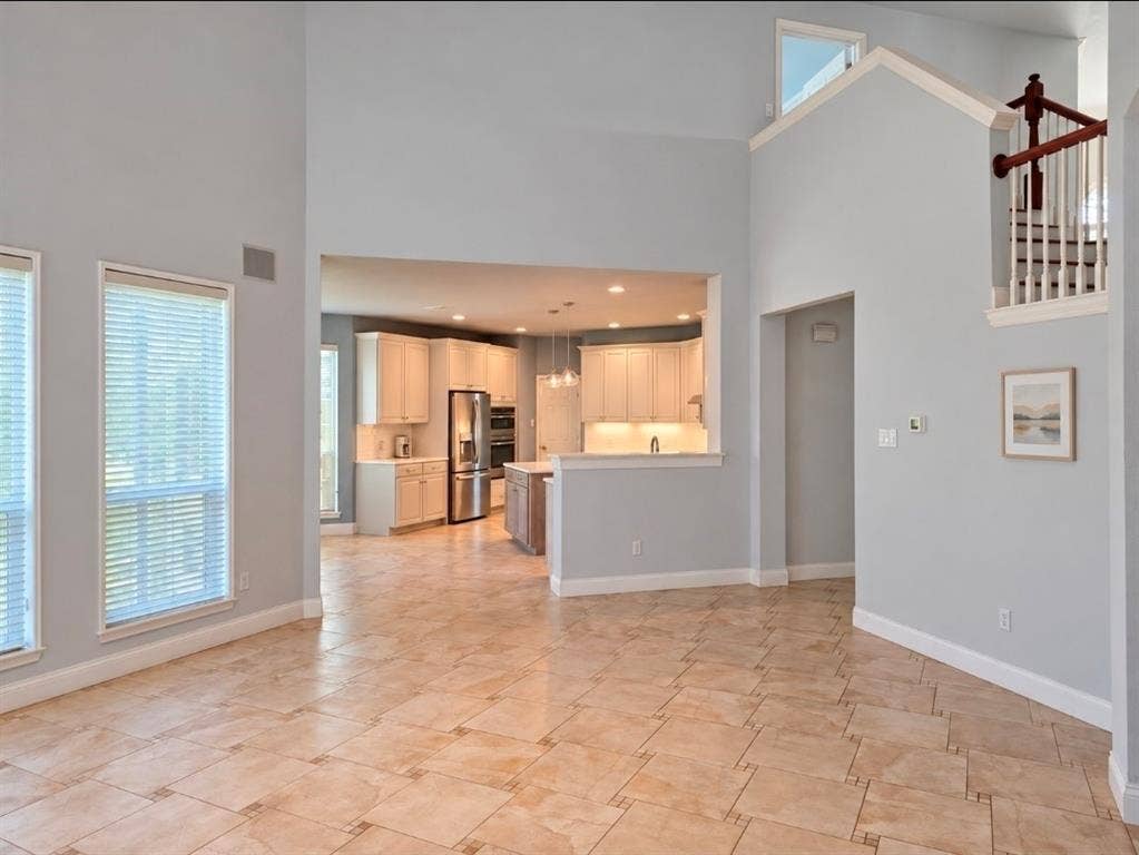 High-ceiling interior featuring tile flooring, a staircase with a wooden handrail, and a kitchen equipped with cabinetry and stainless steel appliances