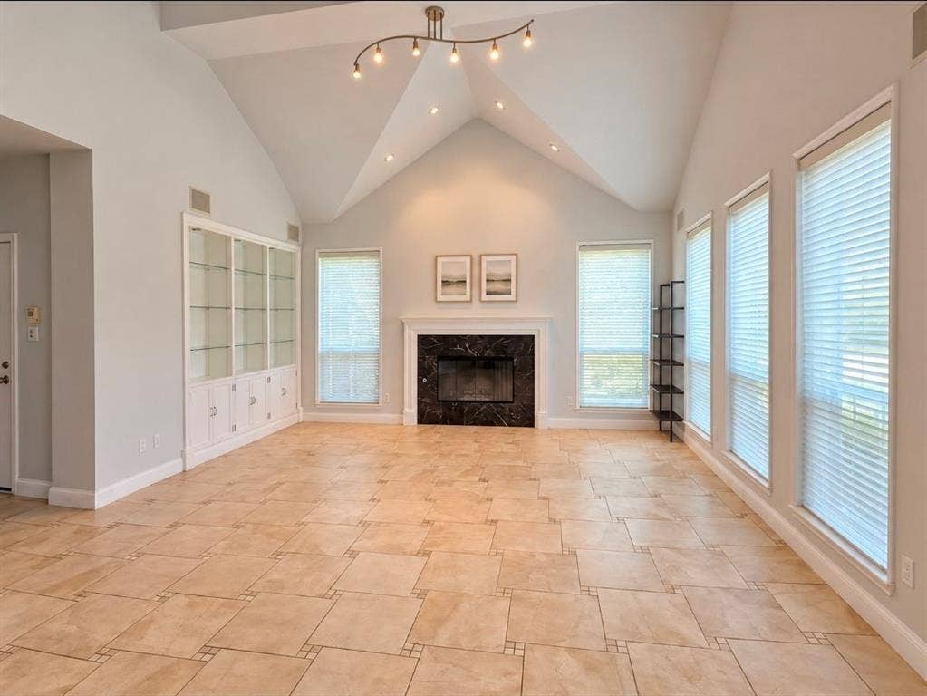 Vaulted ceilings, stone-surround fireplace with mantel, built-in display cabinetry with glass shelving, tile flooring, and multiple tall windows with blinds