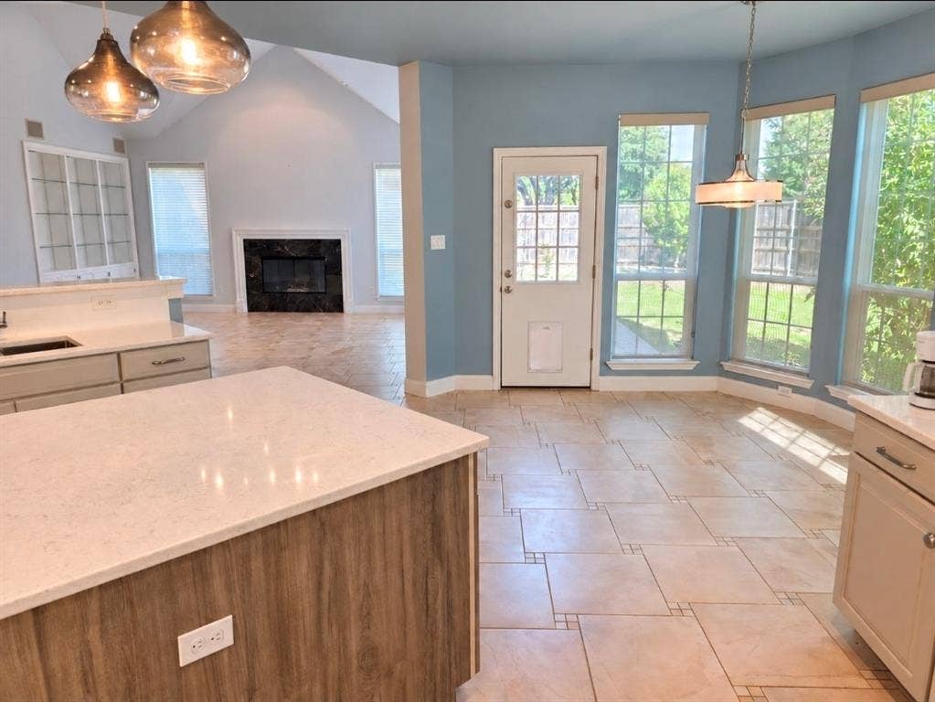Kitchen area featuring a center island with light-toned stone countertop, ceramic tile flooring, built-in display cabinetry, vaulted ceiling, and fireplace with a dark stone surround