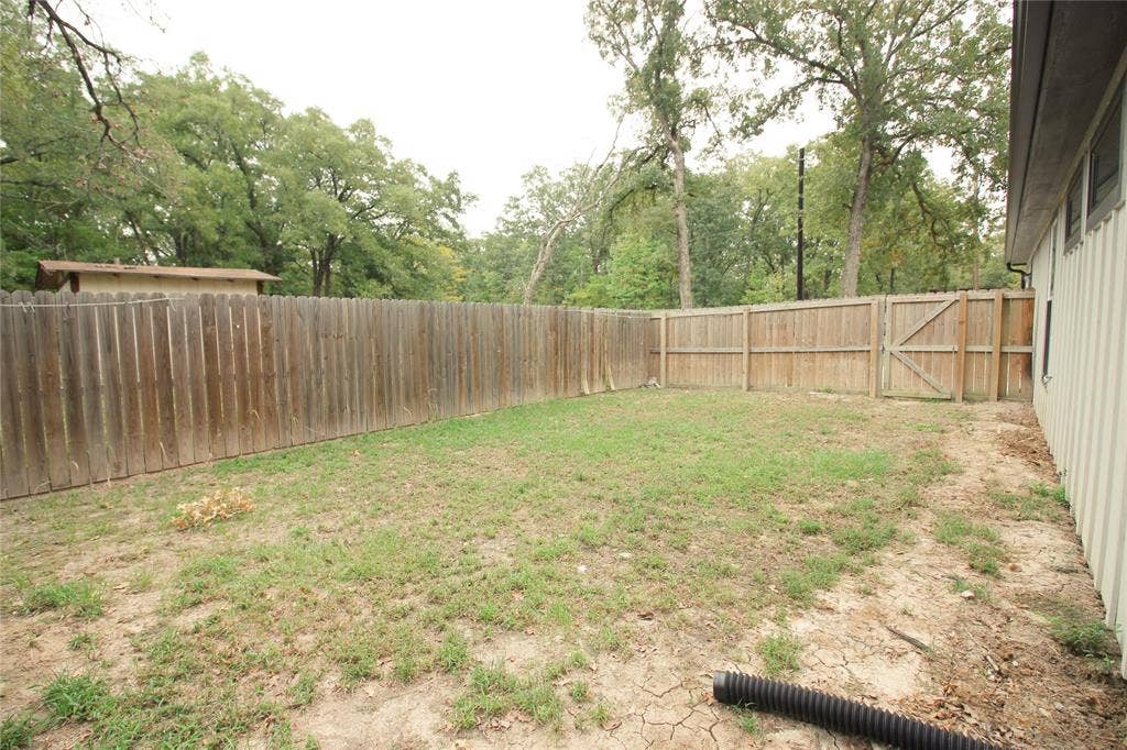 Rear yard enclosed by a tall wood privacy fence with a gated entry, featuring mature trees and vertical siding on the exterior wall