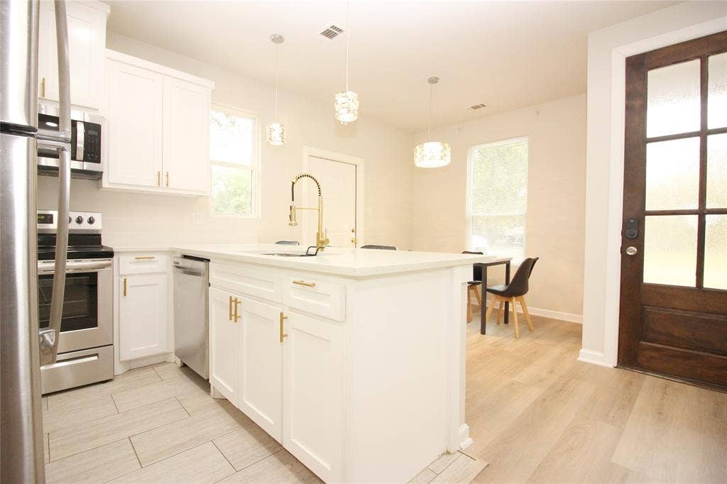 Kitchen featuring white cabinetry with gold-tone hardware, a center island with a quartz countertop and brass-finish faucet, stainless steel appliances, and a wood-finish exterior door with glass panels