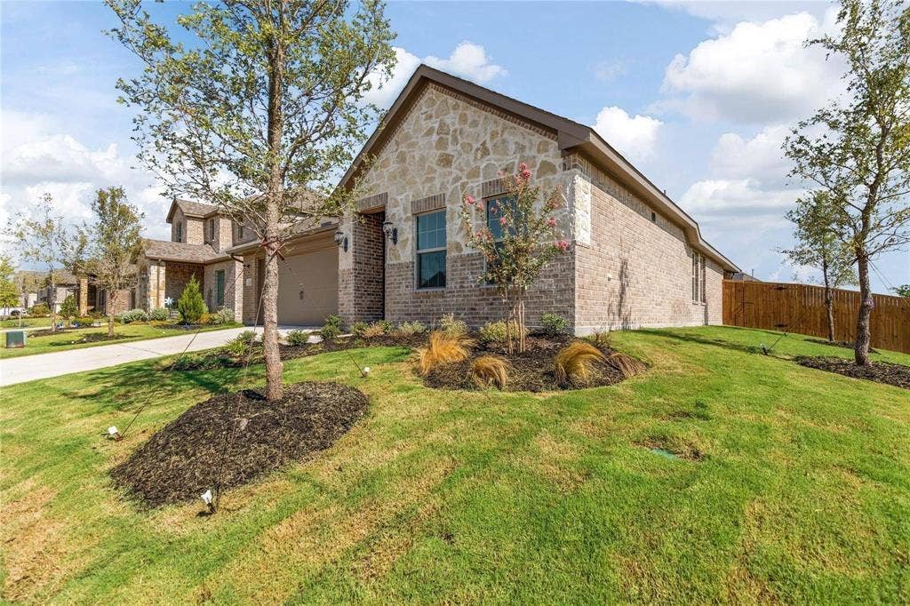 1253 Carinna Drive, Anna, TX 75409 Single-story residence featuring a combination stone and brick facade, gabled roofline, attached multi-car garage, and landscaped front lawn with mulch beds