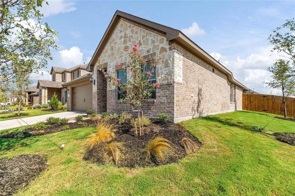 1253 Carinna Drive, Anna, TX 75409 Single-story exterior featuring a combination of brick and stone veneer, a gable roofline, an attached multi-car garage, and landscaped front lawn with mulch beds