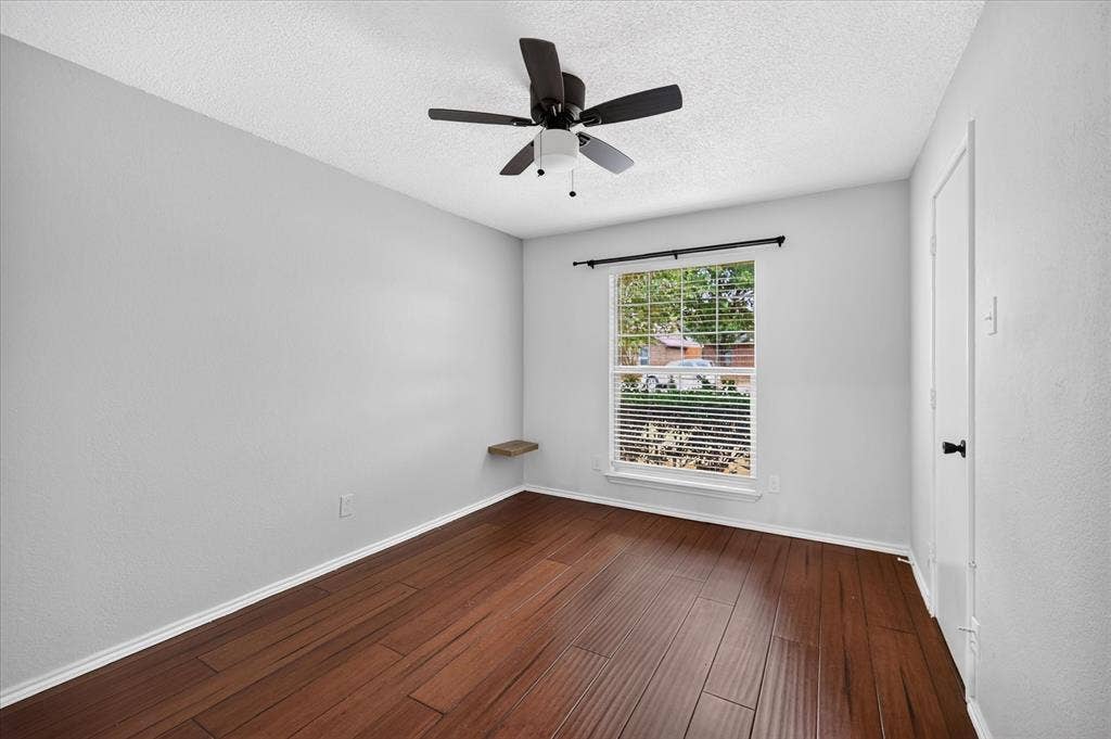 Room featuring wood-finish flooring, a ceiling fan with integrated lighting, a single window with curtain hardware, and a wall-mounted floating shelf