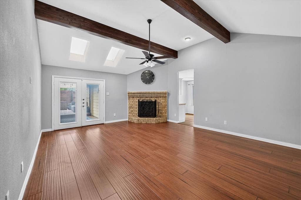 Vaulted ceiling with exposed wood beams, wood-finish flooring, brick corner fireplace, dual skylights, and double-glass paneled exterior doors