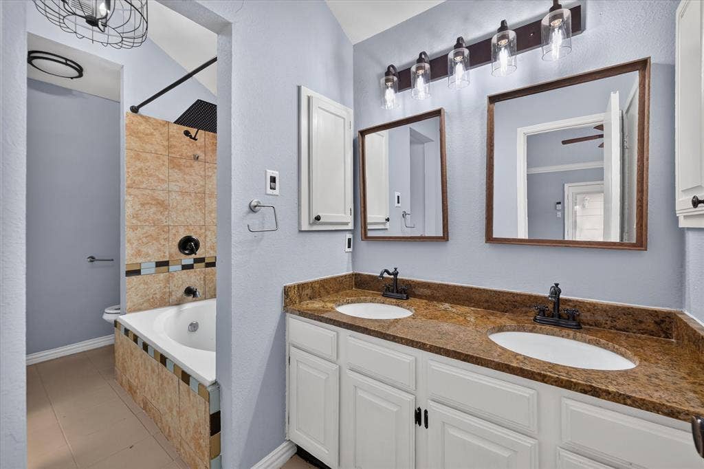 Dual-sink vanity with stone countertop and matte black fixtures, custom wall-mounted mirrors, and soaking tub featuring stone-tile surround