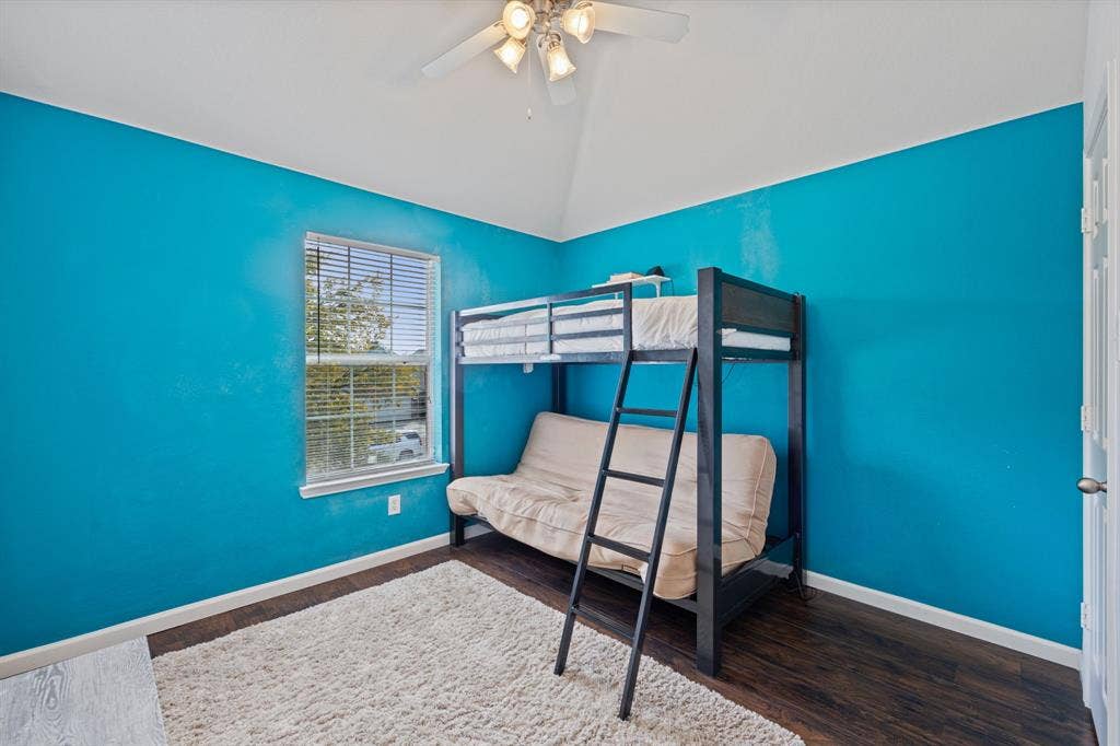 861 Crockett Drive, Lavon, TX 75166 A vibrant bedroom featuring striking turquoise accent walls, a vaulted ceiling, and rich wood-look flooring. A window with blinds brings in natural light while maintaining privacy, and the contrasting white trim adds a crisp, clean finish. The distinctive wall color creates a lively atmosphere, making this room an inviting space with plenty of personality.