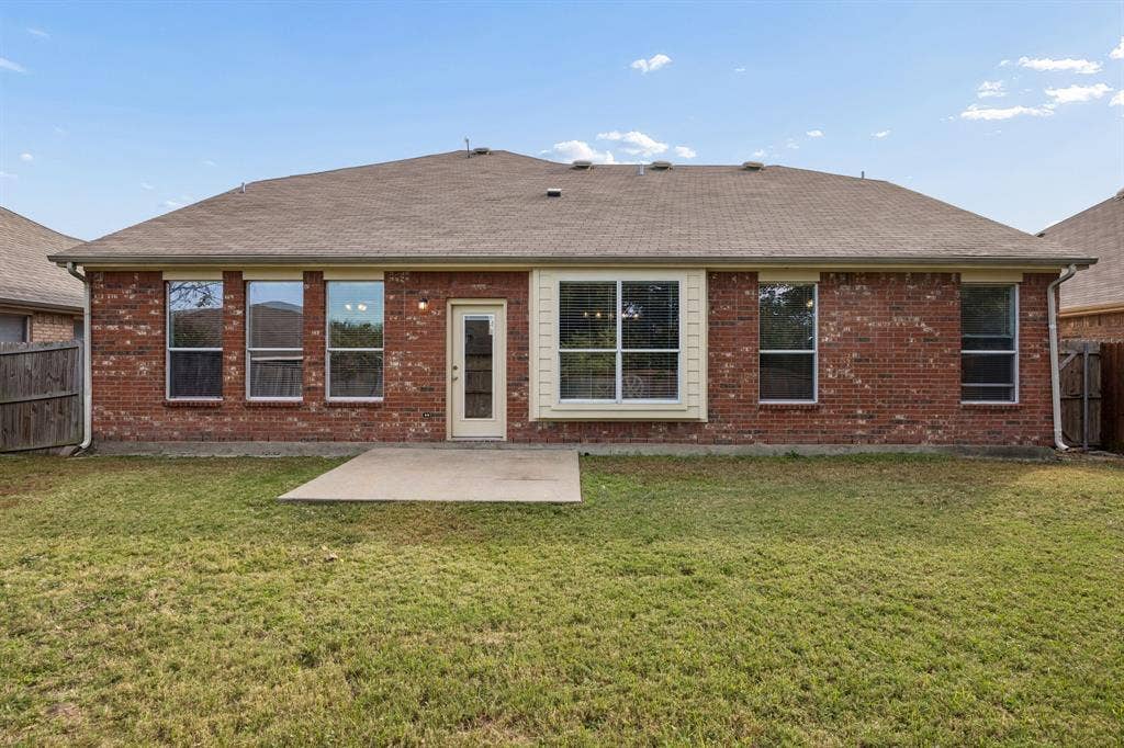 861 Crockett Drive, Lavon, TX 75166 A full view of the home's rear elevation highlights the traditional brick exterior, complementary siding accents, and well-positioned windows that bring natural light into the interior. The fenced backyard features an open grassy lawn and a concrete patio, providing a functional outdoor space with potential for relaxing, entertaining, or creating a garden area.