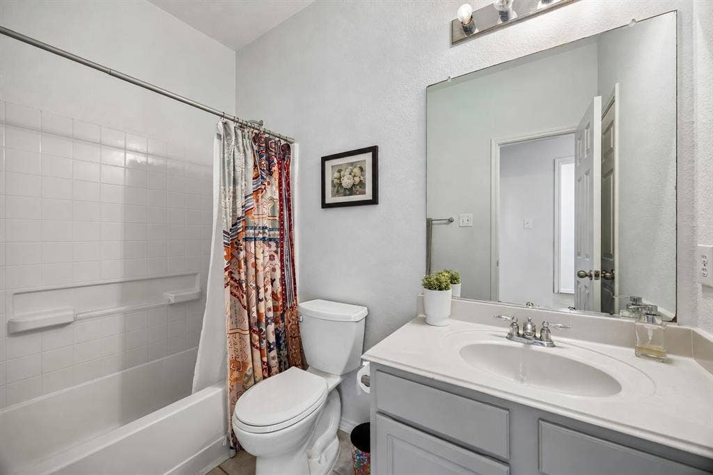 861 Crockett Drive, Lavon, TX 75166 This full bathroom on the first floor features a tub and shower combination with white tile surround, a single-sink vanity, and a large wall mirror with bright vanity lighting. Gray cabinetry complements the light countertop and neutral wall color, while tile flooring provides a durable, easy-maintenance finish. The functional layout offers everyday convenience and a clean, timeless look.