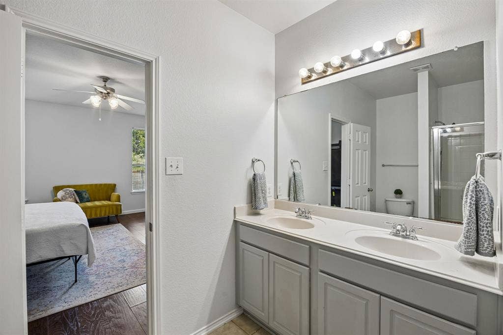 861 Crockett Drive, Lavon, TX 75166 Designed with convenience in mind, this primary bathroom offers a long dual-sink vanity, generous counter space, and a large mirror illuminated by bright vanity lighting. Gray cabinetry adds a subtle contrast to the light countertops and neutral walls, while tile flooring provides a durable, easy-care surface. The layout offers functionality and comfort for daily routines.