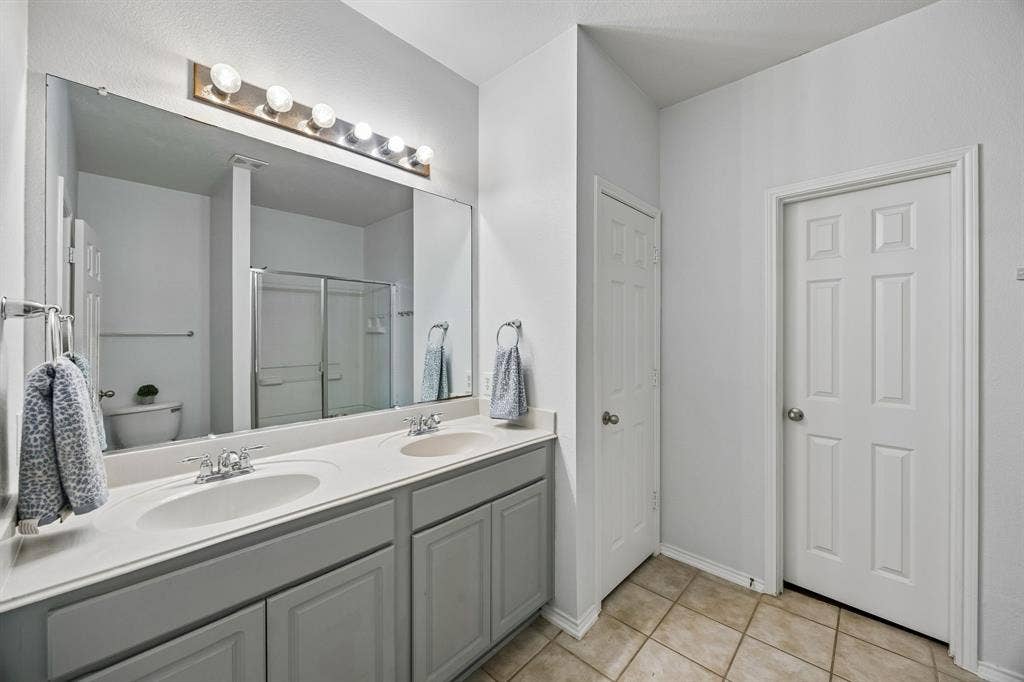 861 Crockett Drive, Lavon, TX 75166 The primary bathroom features an extended dual-sink vanity with ample countertop space, providing added convenience for busy mornings and daily routines. Gray cabinetry, a large wall mirror, and bright vanity lighting contribute to a clean, functional design. Neutral tile flooring complements the surrounding finishes, while the adjoining shower area adds to the room's practicality.