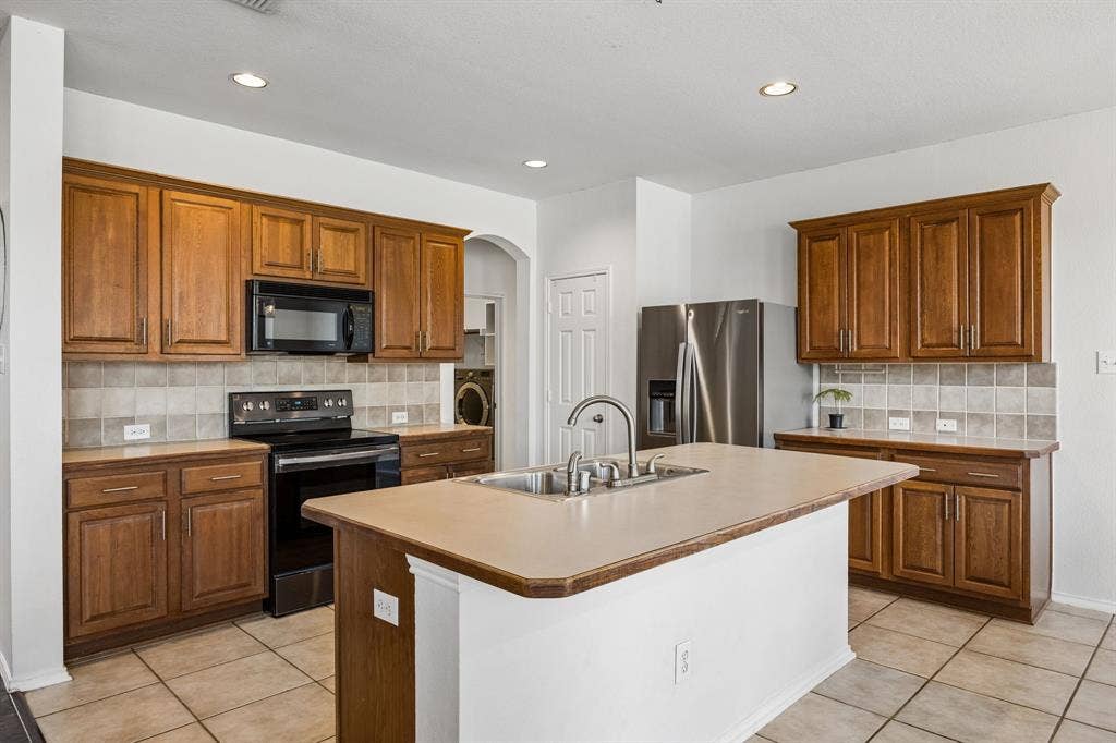 861 Crockett Drive, Lavon, TX 75166 The kitchen offers generous cabinetry, ample countertop space, and a functional layout designed to support everyday cooking and entertaining. Warm wood cabinetry complements the neutral tile backsplash and flooring, creating a classic, inviting aesthetic. A spacious center island with an integrated sink provides additional workspace, while black and stainless steel appliances contribute a polished touch. Recessed lighting keeps the space bright, and the open connection to the adjoining dining and living areas enhances the home's easy flow.