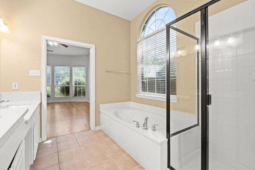 4120 Silverthorne Street, Richardson, TX 75082 Bathroom featuring a soaking tub, arched window, walk-in shower with a black frame, double-sink vanity with solid-surface countertop, and tile flooring