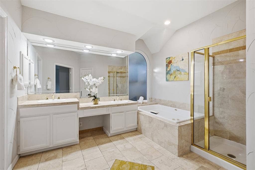 4010 Azure Lane, Addison, TX 75001 A spa-style primary bath with dual sinks, a built-in makeup vanity, a jetted soaking tub, a separate glass-enclosed shower and travertine-look finishes.