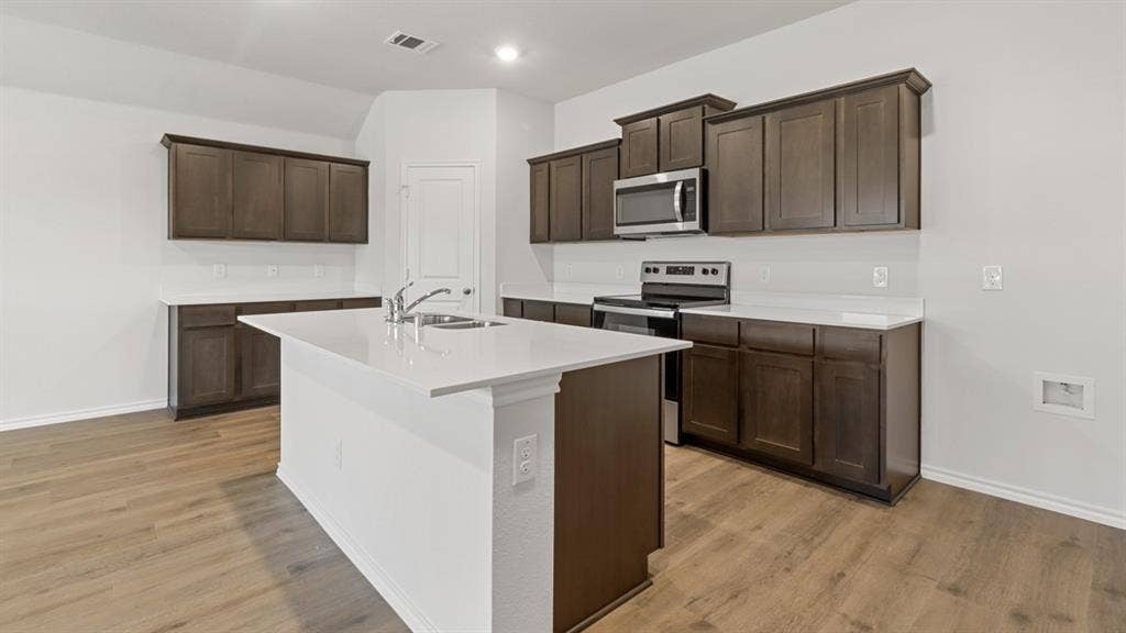 Modern kitchen featuring a central island with a double basin sink, dark wood-finish cabinetry, light-toned countertops, wood-finish flooring, and stainless steel appliances including a range and microwave
