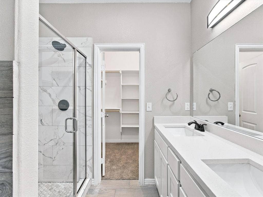 2312 Rogers Avenue, Lancaster, TX 75134 Bathroom featuring a white double-sink vanity with a solid-surface countertop, walk-in shower with stone-tile surround and a frameless glass enclosure, and a built-in walk-in closet with integrated shelving
