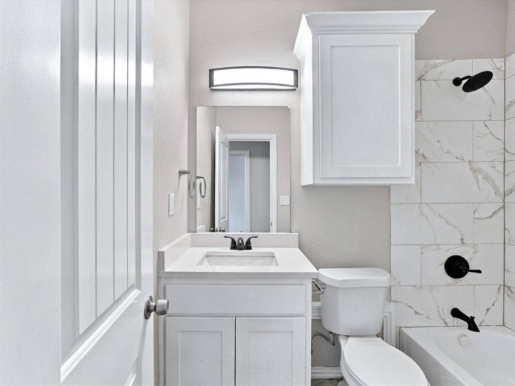 2312 Rogers Avenue, Lancaster, TX 75134 White vanity with a solid-surface countertop and undermount sink, matching wall-mounted cabinetry, tub-and-shower combination with large-format tile surround, and matte black plumbing fixtures