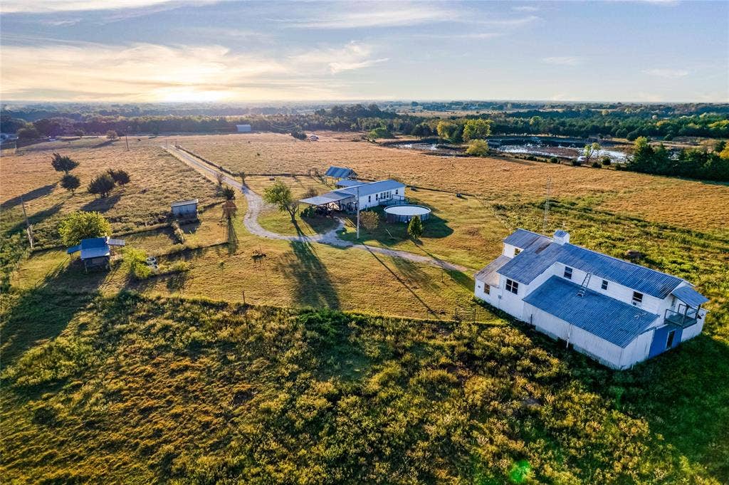 1154 Outlaw Trail, Wolfe City, TX 75496 Expansive acreage featuring multiple structures, metal roofing, a long gravel driveway, and on-site water features