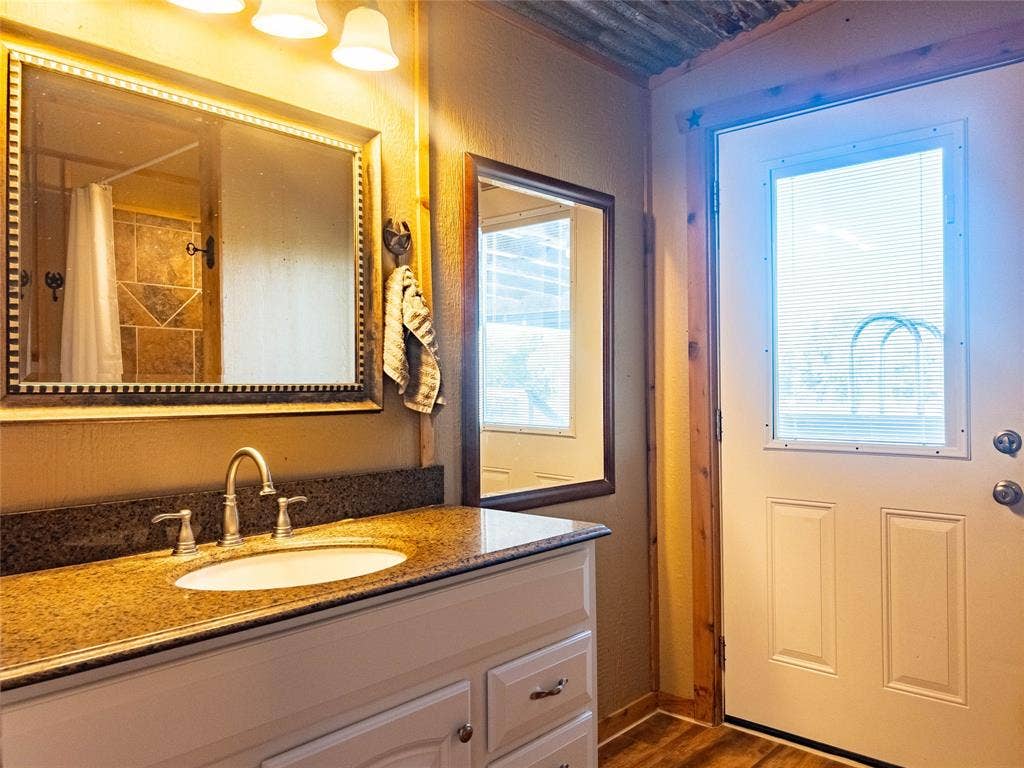 1154 Outlaw Trail, Wolfe City, TX 75496 White cabinetry, stone-finish countertop with integrated sink, three-light vanity fixture, framed wall mirrors, and exterior door with inset glass