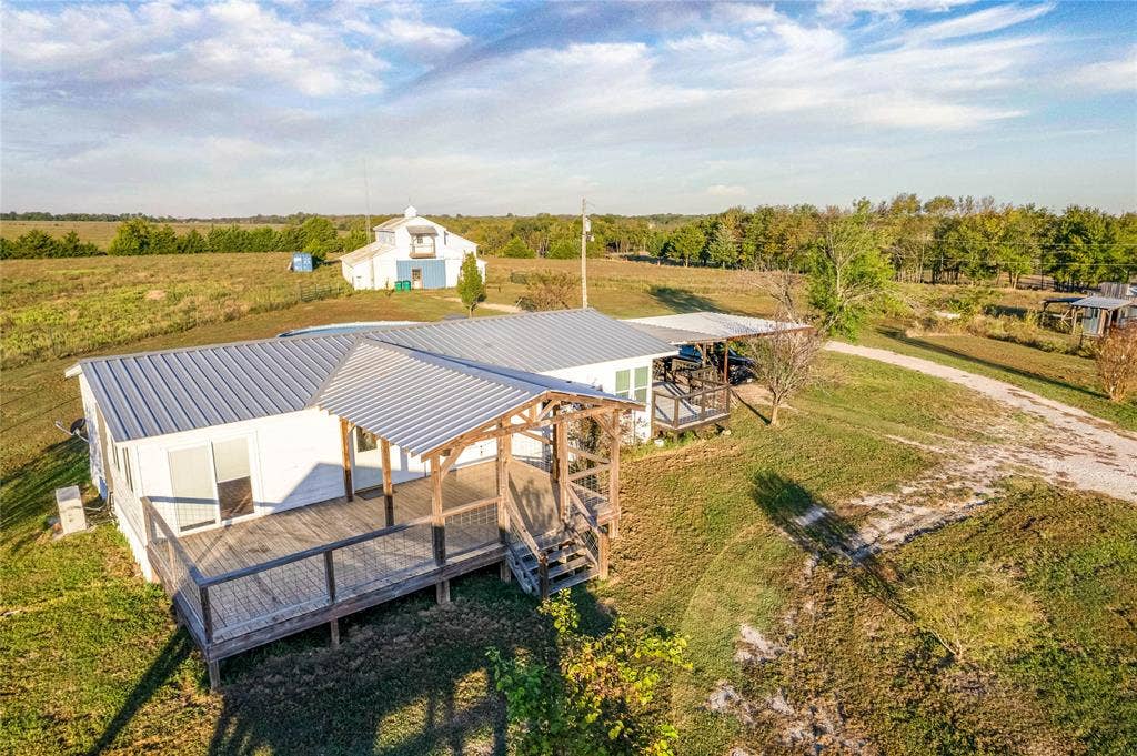 1154 Outlaw Trail, Wolfe City, TX 75496 Exterior featuring a metal roof, wooden deck with railing, and attached covered parking structure situated on expansive acreage