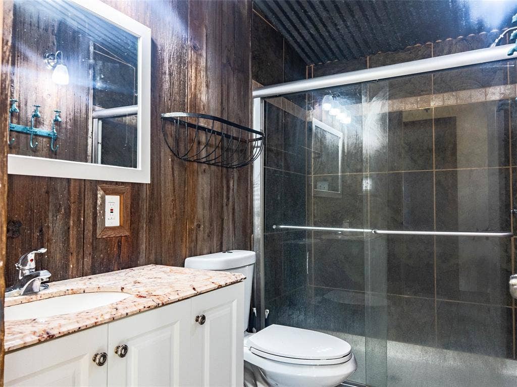 1154 Outlaw Trail, Wolfe City, TX 75496 Bathroom featuring wood-paneled walls, a vanity with a light-toned stone countertop, a glass-enclosed shower with dark tile surround, and a metal corrugated ceiling