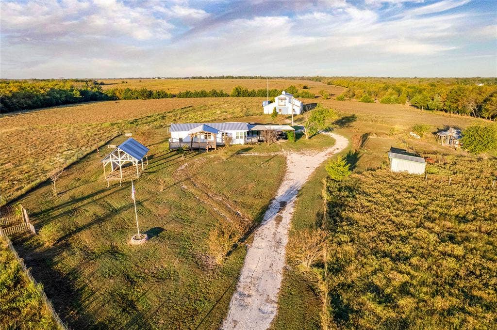 1154 Outlaw Trail, Wolfe City, TX 75496 Aerial perspective of a multi-acre property featuring a single-story residence, a large metal-roofed barn, an open-sided pavilion, and a gravel driveway