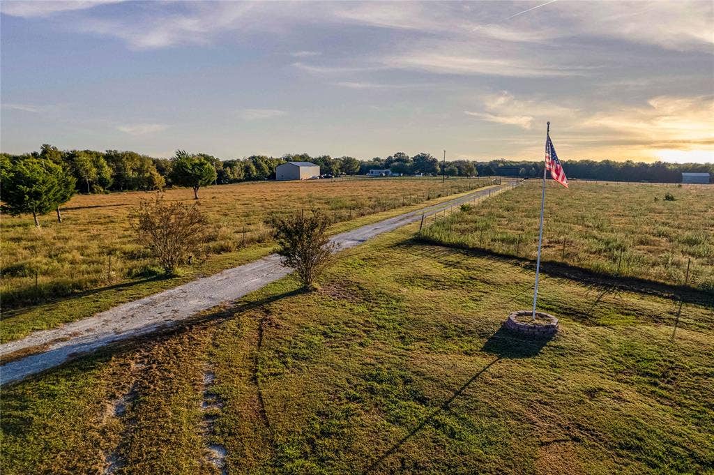 1154 Outlaw Trail, Wolfe City, TX 75496 Expansive acreage featuring a gravel driveway, perimeter wire fencing, a flagpole with a masonry base, and a detached metal outbuilding