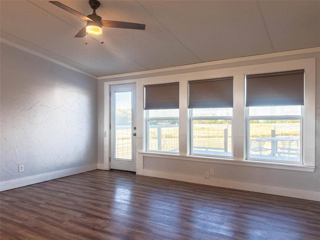 1154 Outlaw Trail, Wolfe City, TX 75496 Room featuring wood-finish flooring, textured accent wall, ceiling fan with integrated lighting, and a bank of three large windows providing natural illumination