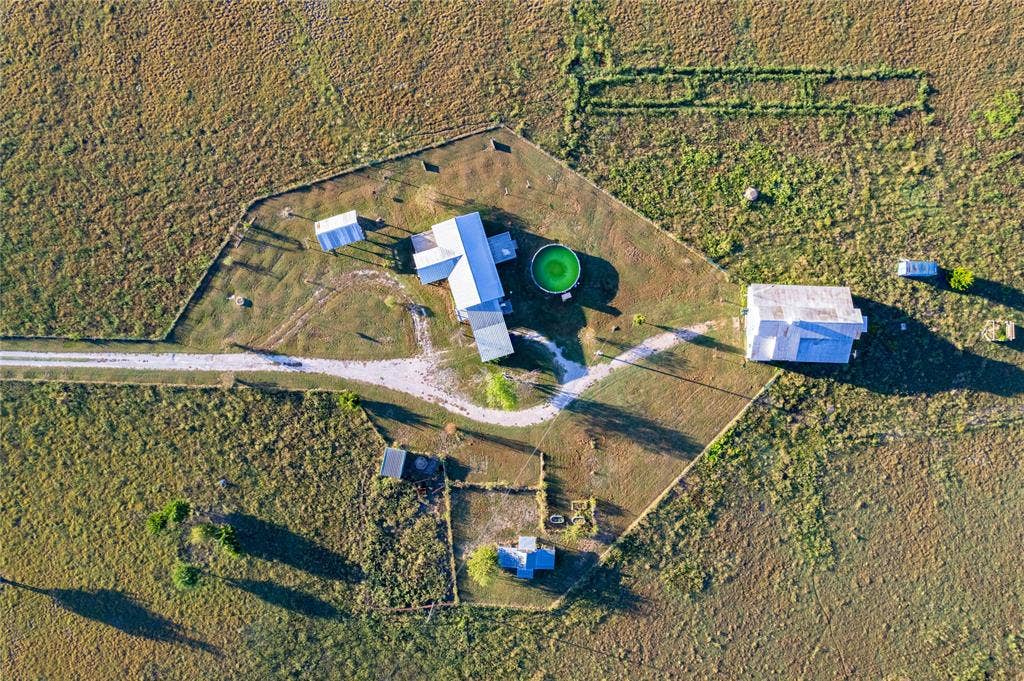 1154 Outlaw Trail, Wolfe City, TX 75496 Aerial perspective of a multi-structure property featuring a main residence, additional outbuildings, a circular pool, and a gravel driveway, all enclosed within a fenced perimeter