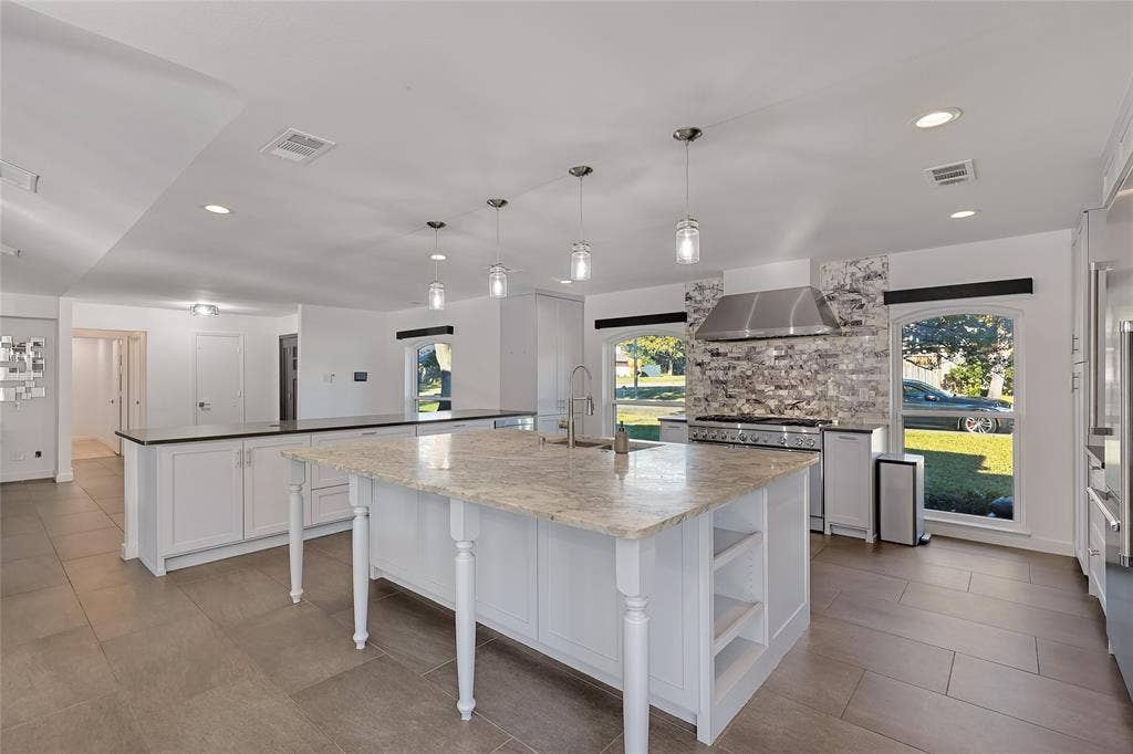 Kitchen featuring a large central island with stone countertop, stone-tiled backsplash, stainless steel range hood, pendant lighting, and oversized tile flooring