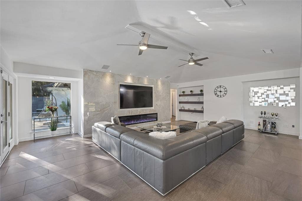 Living space featuring large-format ceramic tile flooring, a vaulted ceiling with dual ceiling fans, a stone-finish fireplace surround, an integrated linear electric fireplace, and built-in shelving