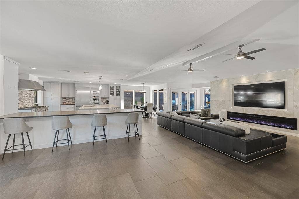 Open-concept interior featuring large-format tile flooring, a chef's kitchen with a stone-top center island, a linear electric fireplace set within a floor-to-ceiling stone surround, and dual ceiling fans