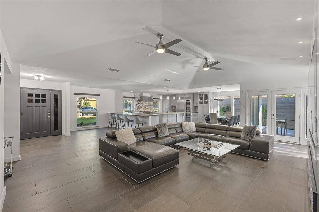 Open-concept living space featuring vaulted ceilings, large-format tile flooring, dual ceiling fans, a kitchen island with stone-finish backsplash, and multi-light glass paneled exterior doors
