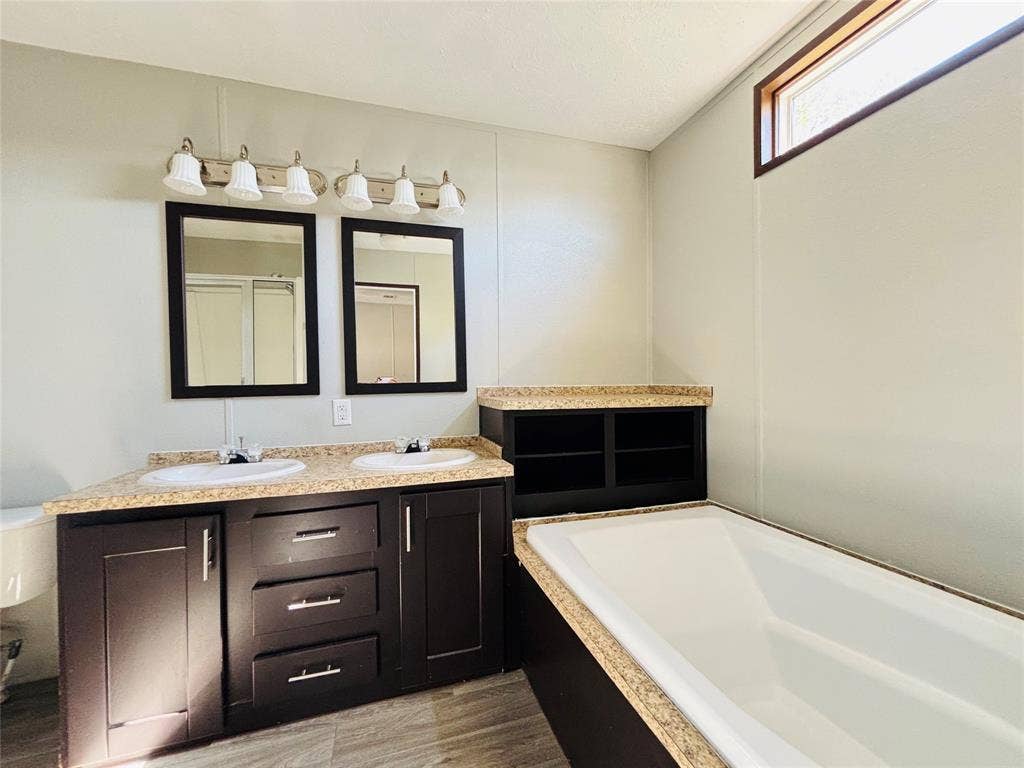 14311 Skyfrost Drive, #116, Dallas, TX 75253 Dual vanity featuring dark cabinetry, stone-finish countertops, undermount sinks, and two wall-mounted mirrors, accompanied by a built-in soaking tub with a matching surround and horizontal clerestory window
