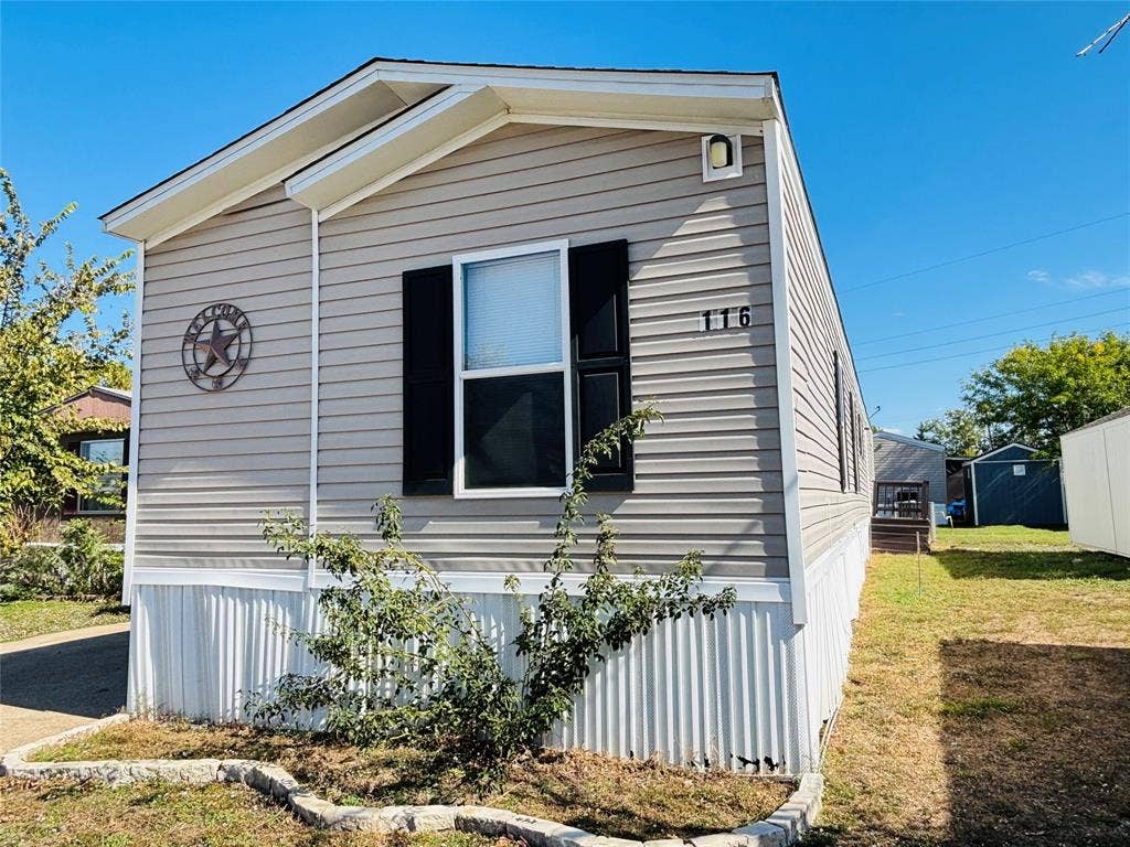 14311 Skyfrost Drive, #116, Dallas, TX 75253 Exterior featuring light-tone horizontal vinyl siding, black window shutters, a white corrugated metal skirting, and a stone-bordered landscape edging