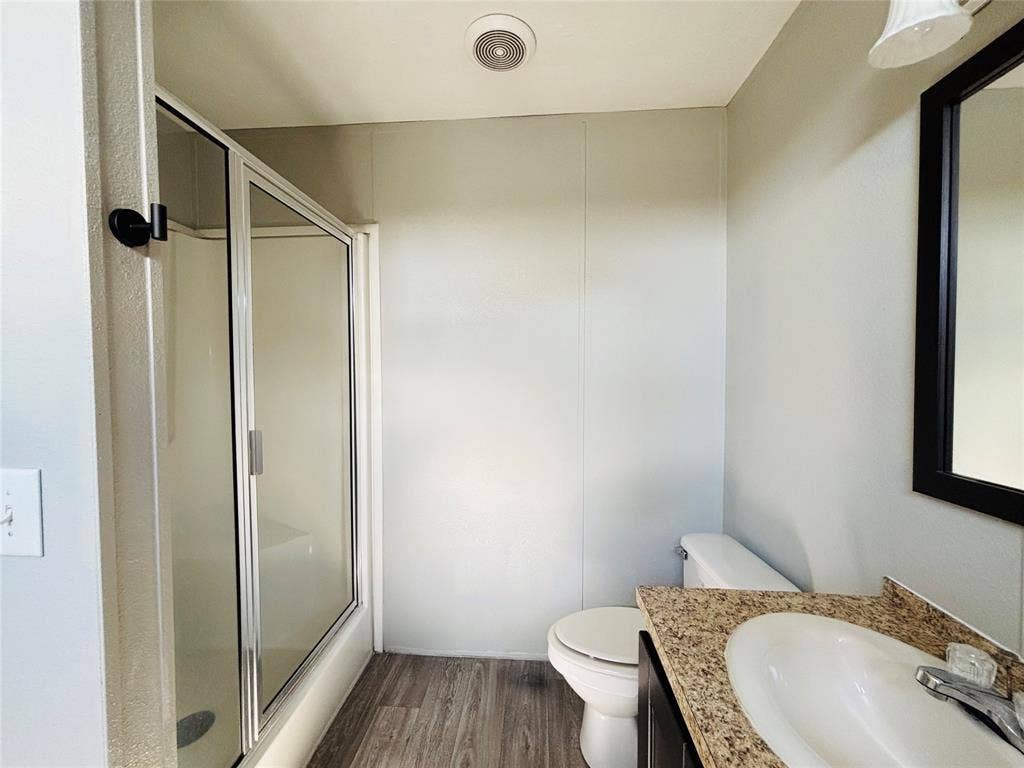 14311 Skyfrost Drive, #116, Dallas, TX 75253 Bathroom featuring a glass-enclosed shower, stone-finish countertop with an integrated basin, wood-finish flooring, and a framed vanity mirror