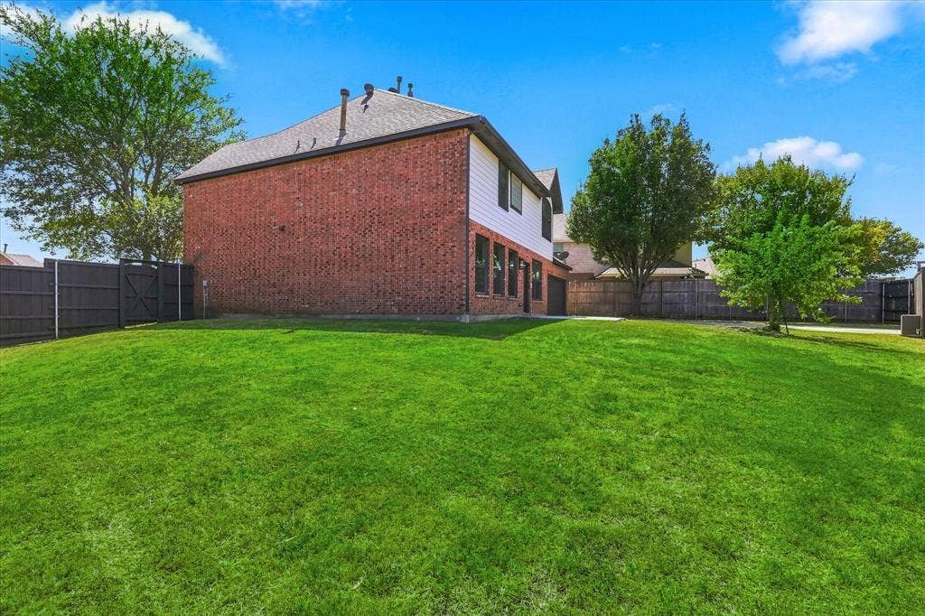 4421 Red Barn, Richardson, TX 75082 Rear exterior featuring red brick and white horizontal siding, a gabled roof, expansive grass lawn, and full-perimeter wood privacy fencing